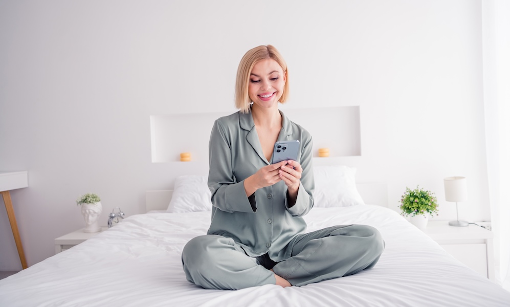 Full body length photo of young lady blonde hair model sitting bed white laundry texting sms on iphone her boyfriend buy sweets indoors
