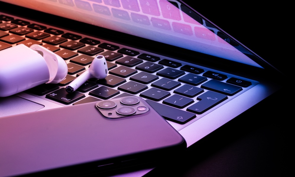 a laptop half closed in the dark with a phone and wireless earphones glowing