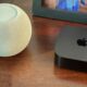 A white Apple HomePod mini sitting next to an Apple TV 4K on a wooden table, representing a smart home hub setup.