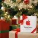 A white AppleCare+ package tied with a red ribbon sits prominently among other wrapped gifts under a brightly lit and decorated Christmas tree, with stockings hanging on a fireplace in the background.