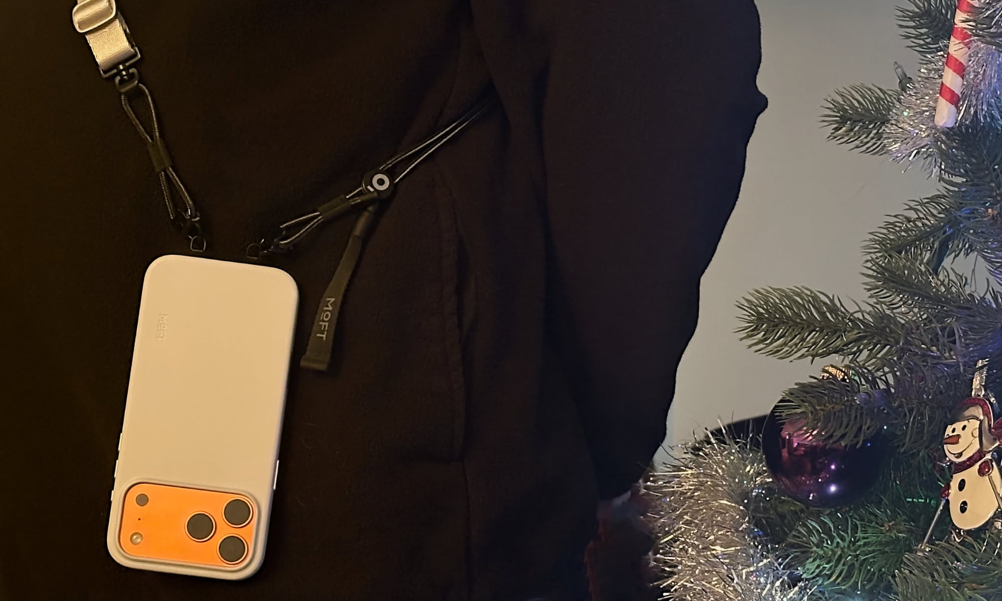 Person standing beside Christmas tree wearing a MOFT Everywhere Lanyard with an orange iPhone 17 Pro Max attached.