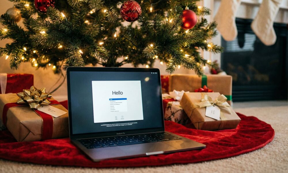 An open MacBook Pro displaying the "Hello" setup screen, sitting on a red tree skirt under a decorated Christmas tree with wrapped gifts.