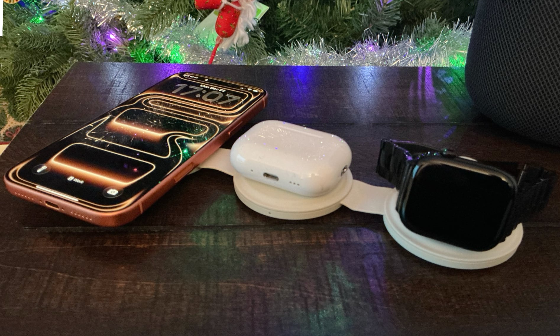 Satechi OntheGo 3-in-1 Travel Charger on a table by a Christmas tree with an orange iPhone 17 Pro Max, AirPods, and Apple watch charging on it.