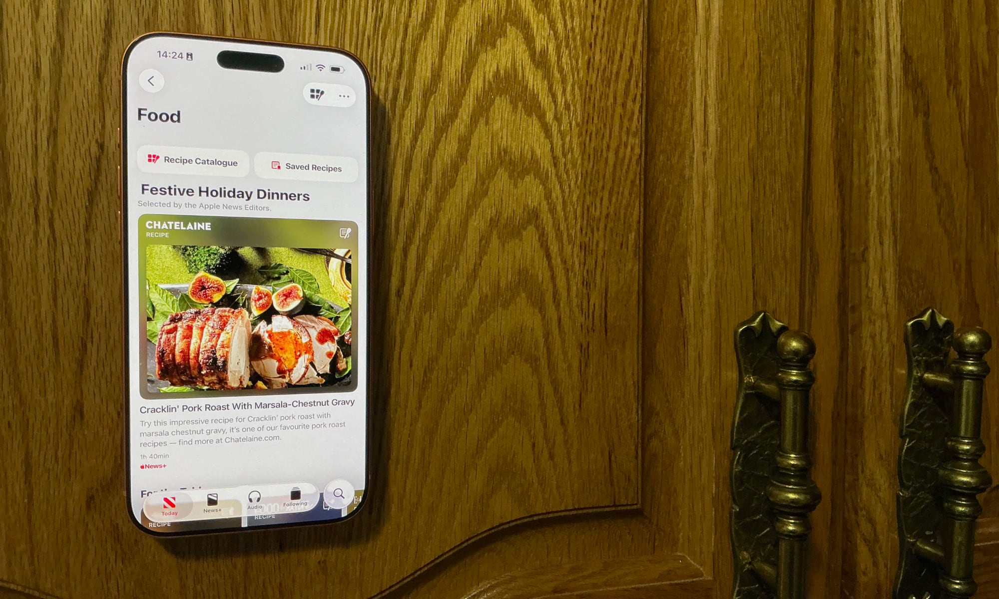 Orange iPhone 17 Pro Max attached to Smartish Mountaineer showing Apple News Food articles on wooden kitchen cabinet door.