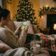 Woman relaxing on a couch playing an iPhone game in a Christmas living room setting with a cat in her lap, a crackling fireplace in the background, and a cup of hot cocoa by her side.