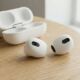 A concept of white 'AirPods Ultra' earbuds without stems, featuring small circular lenses for IR cameras, resting on a table next to an open charging case.