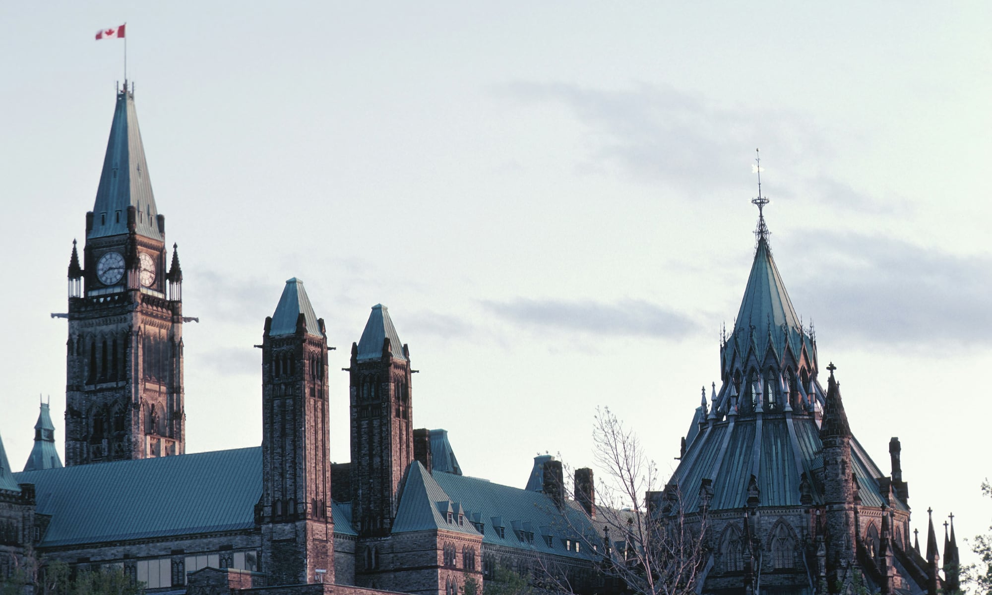 A wide-angle, professional photograph of the Centre Block of the Canadian Parliament buildings in Ottawa. The Gothic Revival architecture is featured prominently, with the Peace Tower rising in the center and the Canadian flag flying from the spire against a clear sky.