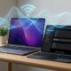 A MacBook Pro and iPad Pro on a wooden desk with glowing blue Wi-Fi signal waves and a signal icon floating above them, representing a high-speed wireless connection.