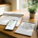 A sunlit wooden kitchen table with a 2026 calendar open to January, an iPhone displaying a goal-tracking app, and a handwritten list of New Year's resolutions.