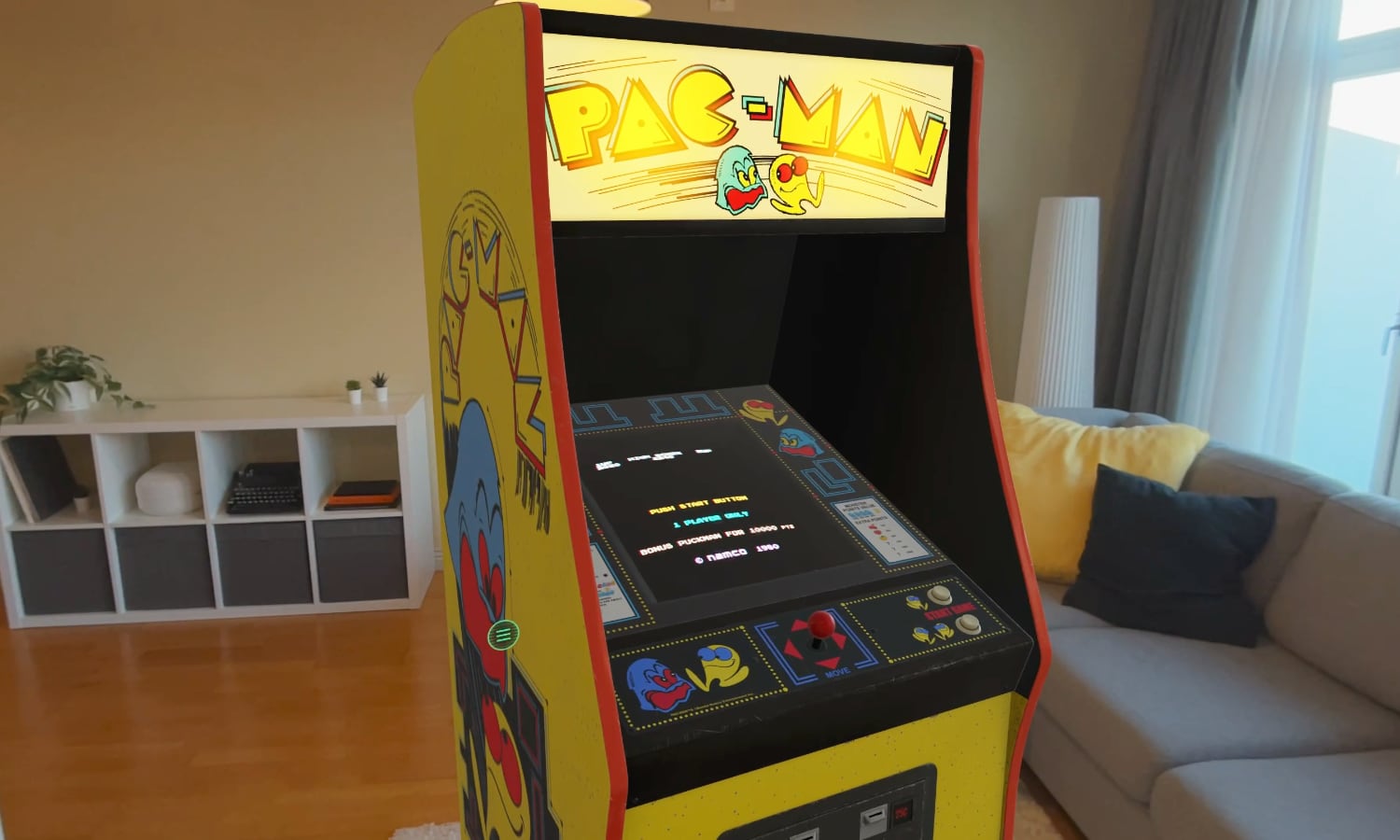 A high-fidelity digital model of the legendary yellow Pac-Man arcade machine, rendered as spatial furniture within a real-world room via the Apple Vision Pro. The image shows the cabinet's original character art, the classic red joystick, and a glowing CRT-style screen displaying the Pac-Man maze, complete with realistic shadows and lighting.