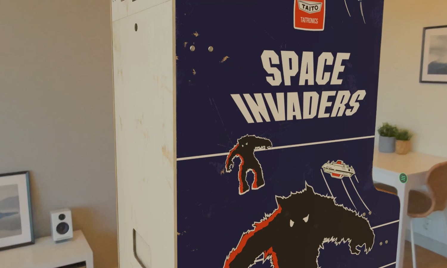 A high-detail virtual replica of the iconic Space Invaders arcade machine, viewed as a piece of "spatial furniture" in the Retrocade app. The cabinet displays the legendary alien invaders on its screen and features the original colorful cabinet graphics, placed within a modern room using the Apple Vision Pro&rsquo;s passthrough technology.
