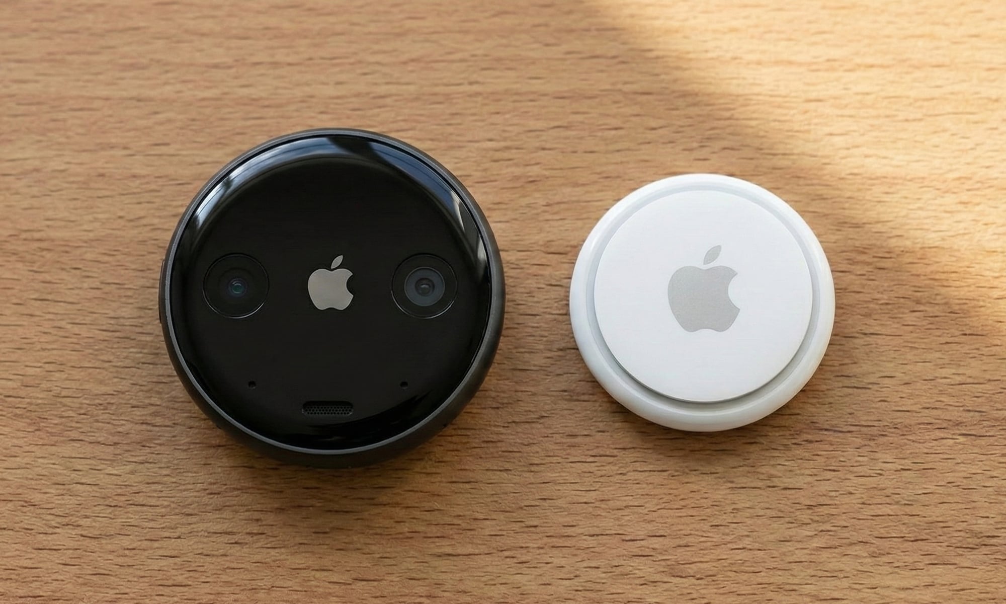 A top-down photograph comparing the size of a conceptual black Apple AI Pin (left) with a standard white Apple AirTag (right) resting on a wooden desk. The AI pin features two cameras and an Apple logo on its face and is noticeably thicker than the AirTag beside it.