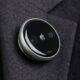 A concept design of a circular Apple AI pin attached to a charcoal grey suit lapel. The device features a polished aluminum rim, a black glass face with a small silver Apple logo in the center, and two visible camera lenses on either side. A physical button is visible on the side of the thin, disc-shaped wearable.