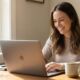 A happy woman smiling while using her responsive MacBook Pro at a bright home office desk with a coffee mug.