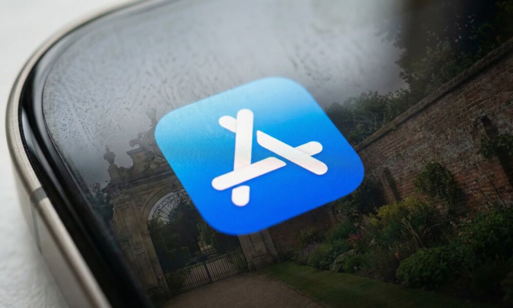 A macro photograph of an iPhone screen displaying the glowing App Store icon. Reflected on the glass surface is a large stone gate and high brick walls enclosing a garden, illustrating the "walled garden" metaphor central to Apple's antitrust challenges.