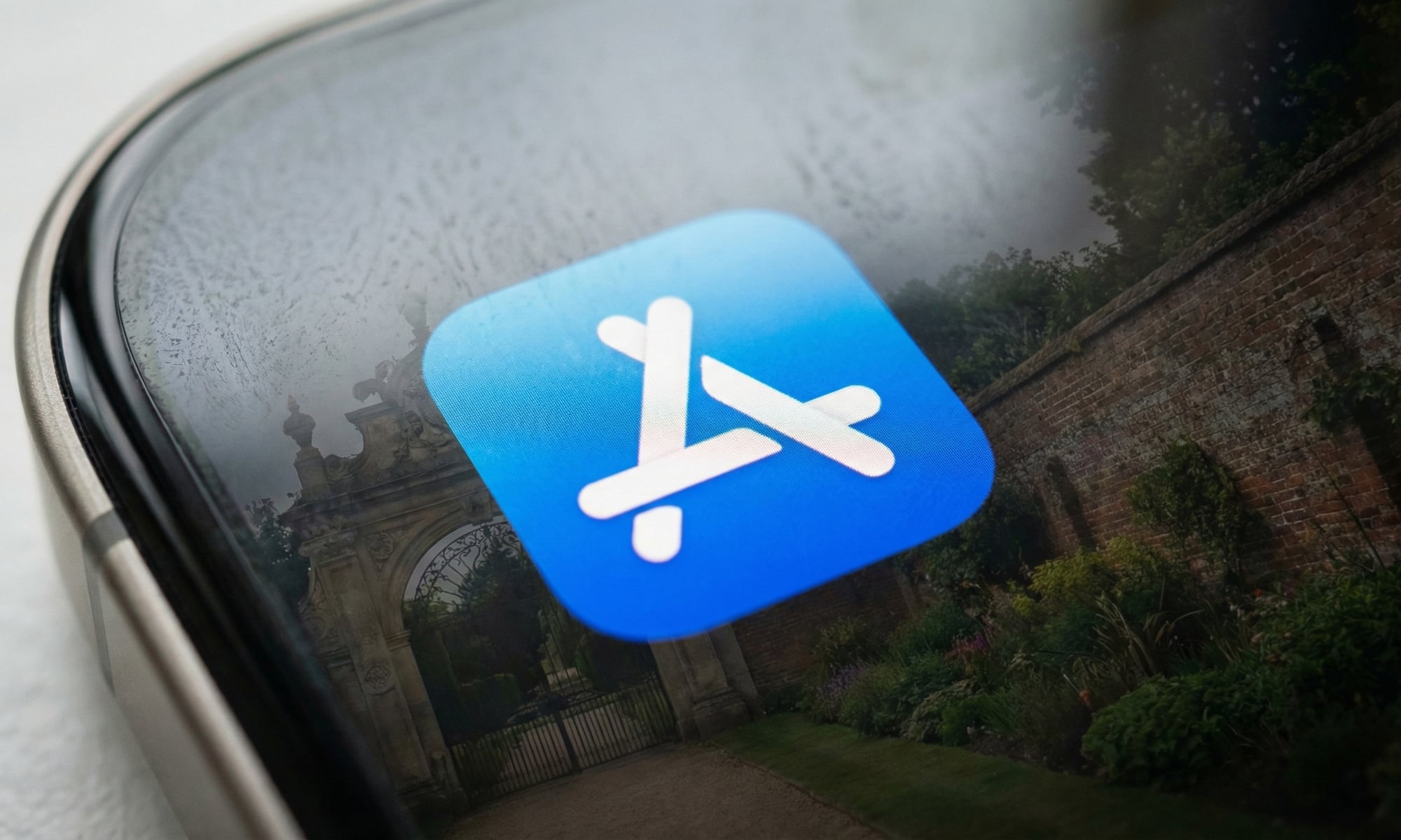 A macro photograph of an iPhone screen displaying the glowing App Store icon. Reflected on the glass surface is a large stone gate and high brick walls enclosing a garden, illustrating the "walled garden" metaphor central to Apple's antitrust challenges.