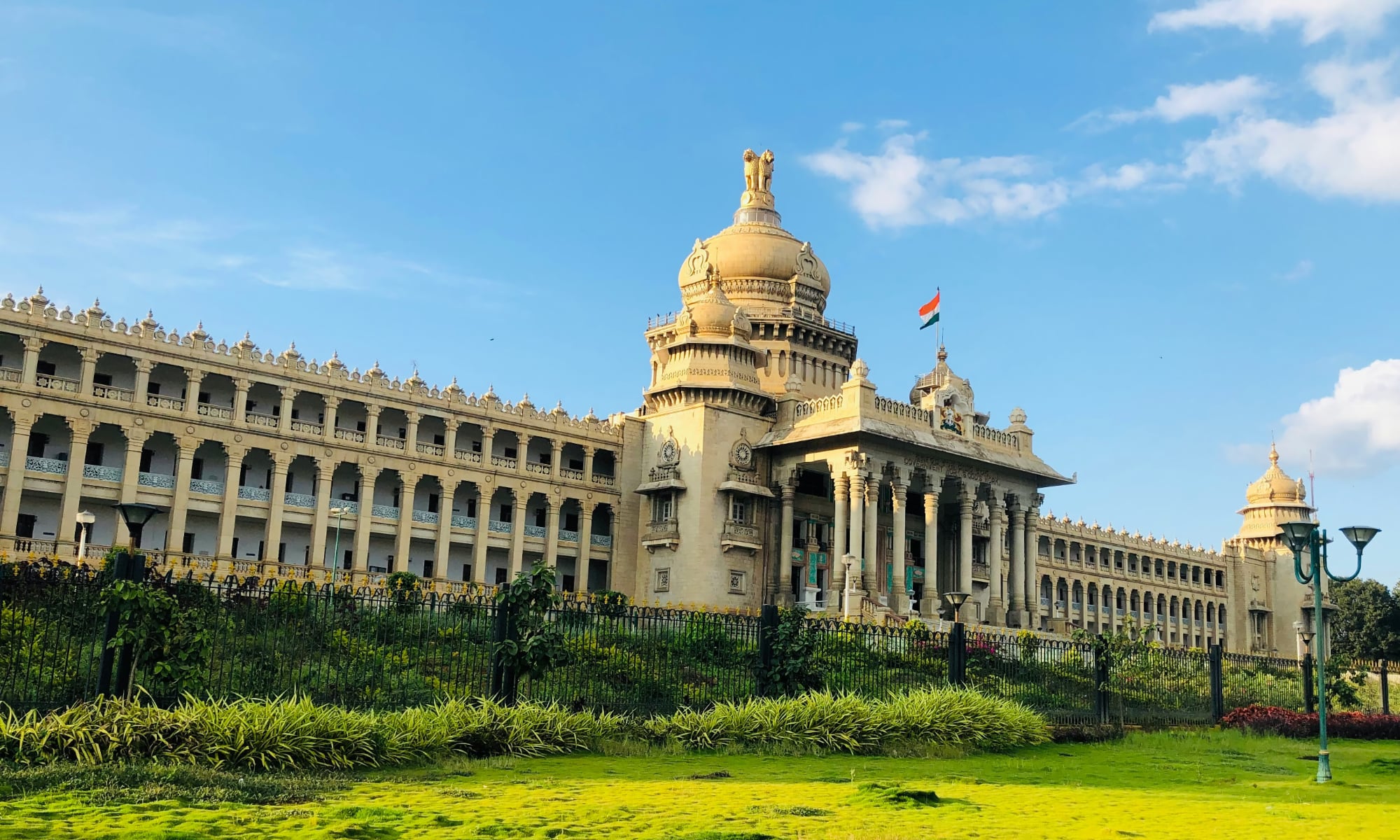 A wide exterior view of the Vidhana Soudha in Bengaluru, a massive Neo-Dravidian granite building featuring a grand central dome topped by the National Emblem of India, towering columns, and a wide flight of stairs leading to the entrance.