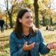 A smiling woman in a denim jacket wearing AirPods Pro earbuds and gesturing while having a conversation with a friend in a sunny autumn park, demonstrating the Conversation Awareness feature.