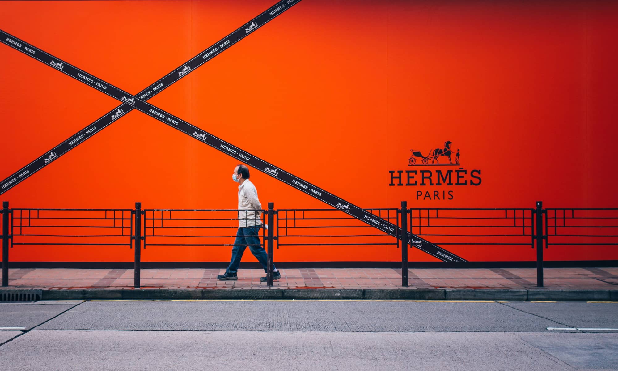 A man wearing a face mask walks past a massive, vibrant orange Herm&egrave;s Paris billboard in Tsim Sha Tsui, Hong Kong. The billboard features the iconic horse-and-carriage logo and crisscrossing black ribbons with "Herm&egrave;s - Paris" branding.