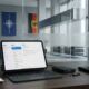 An iPad Pro on a Magic Keyboard and an iPhone resting on a modern wooden desk in a secure office environment. In the blurred background, the NATO and German flags are visible through a glass partition featuring etched security-themed icons.