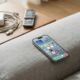 A modern iPhone 17 Pro resting on a grey fabric couch, with an old silver flip phone and a tangled white charging cable sitting on a wooden side table in the background.