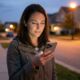A woman standing on a quiet residential sidewalk at dusk, looking down at a modern iPhone with a triple-lens camera system while warm streetlights glow in the background.