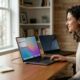 A woman sitting at a bright wooden desk, smiling while using a MacBook Pro running macOS 26. A Windows laptop sits beside it, illustrating the transition from PC to Mac in a modern home office.