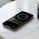 A sleek iPhone 19e concept sitting on a minimalist marble cafe table next to a glass of cold brew, its screen showing a vibrant always-on clock with smooth fluid animations.