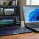 A side-by-side comparison shows the incredibly thin Indigo MacBook Neo editing 4K video in Final Cut Pro next to a significantly thicker, bulky black textured plastic budget PC laptop, highlighting the design and performance gap.