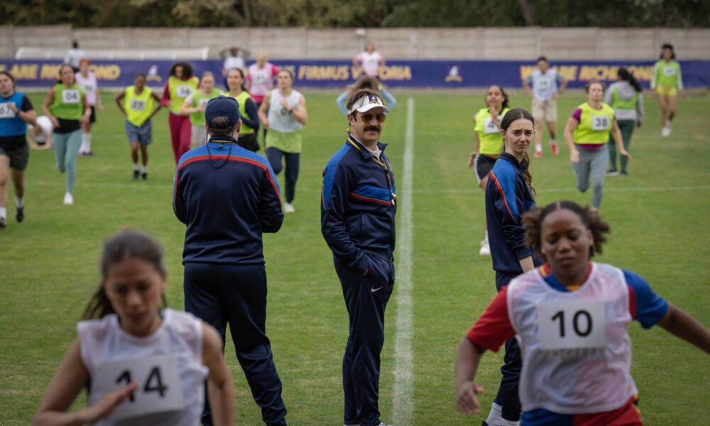 Jason Sudeikis as Ted Lasso standing on a busy football pitch.