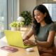 A diverse young professional smiling while working at a modern, sunlit desk with the 'Citrus' MacBook Neo, soft pink iPhone 17e, Apple Watch SE 3, and AirPods 4 arranged together, showcasing a complete and affordable Apple ecosystem in 2026.