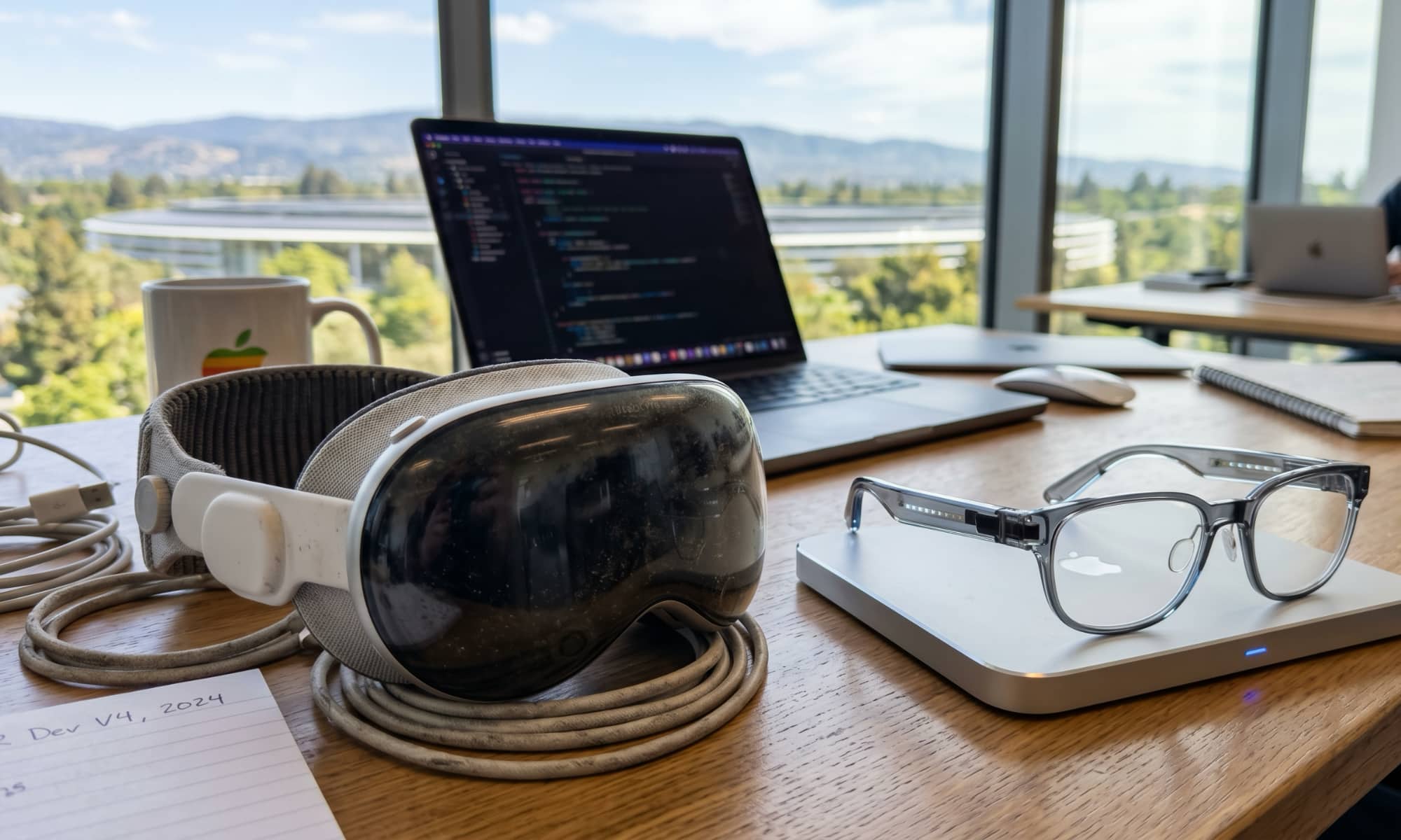 A high-end workspace featuring a dusty Apple Vision Pro M5 headset sitting next to a pair of sleek, prototype-style AR smart glasses on a charging pad.
