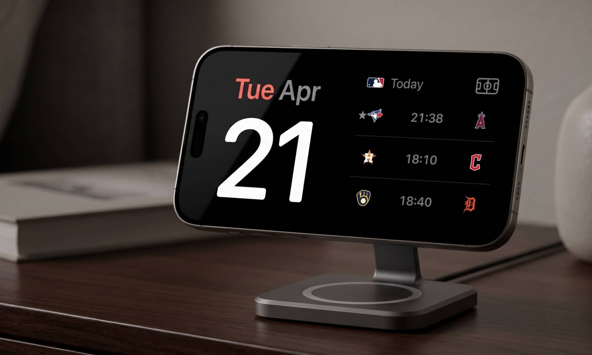 A close-up photograph of an iPhone on a dark wood nightstand, connected to a MagSafe charging stand. The landscape StandBy mode UI is active, displaying "Tue Apr 21" alongside live MLB sports score widgets against a black background.