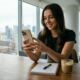 A young woman smiling while using her iPhone at a wooden desk in a modern high-rise office overlooking a city skyline.