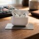 A set of AirPods Pro 3 with the case open, resting on top of a physical titanium Apple Card on a grained wooden desk. The front of the open case clearly displays 'AirPods Pro 3'. The image is a close-up lifestyle shot with natural lighting.