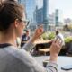 An over-the-shoulder view of a woman wearing minimalist AR smart glasses at an outdoor city cafe patio. She performs a pinch gesture, interacting with holographic user interface icons floating in the air before her. A smartphone rests on the table next to a coffee cup.