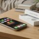 A lifestyle photograph of a newly unboxed iPhone 17 Pro Max on a wooden living room coffee table. The phone shows rows of app icons like Spotify, Signal, and 1Password against a warm living room background.