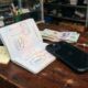 A black iPhone plugged into a charging cable on a rustic wooden desk inside a bustling Vietnamese electronics repair shop. Next to the device lies an open Canadian passport displaying travel stamps from Toronto and Hanoi, along with Vietnamese Dong banknotes and a yellow sticky note reading 