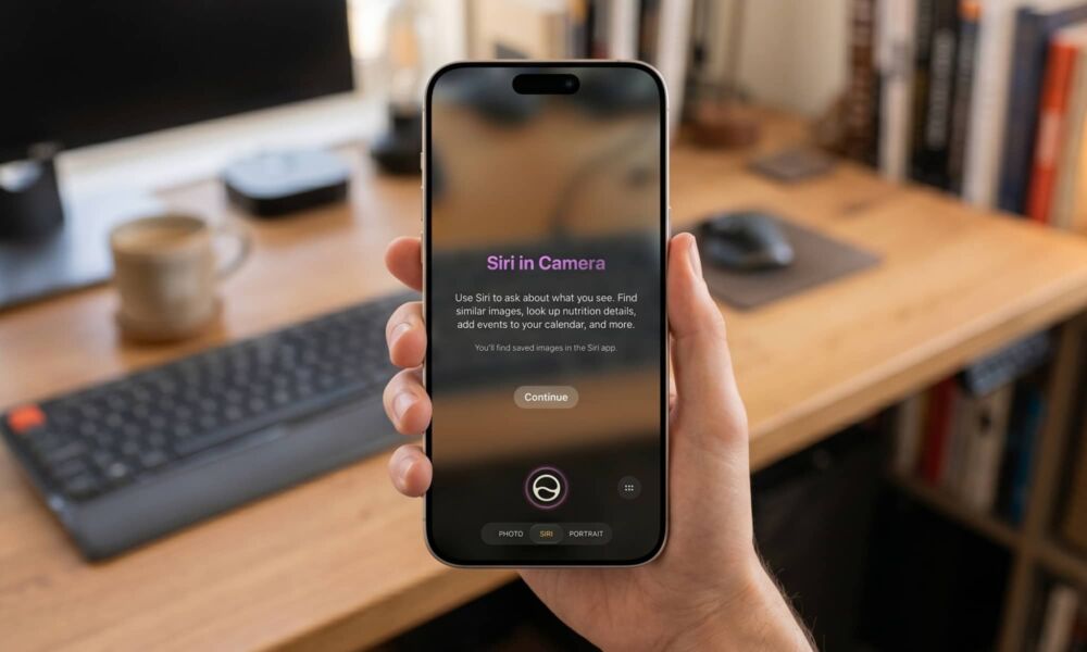 A first-person close-up of a hand holding an iPhone 17 Max in a bright, modern home office with a blurred wooden desk background. The iPhone screen displays the new iOS 27 "Siri in Camera" interface, showcasing the native Visual Intelligence onboarding text and the dedicated Siri camera shooting mode.