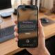 A first-person close-up of a hand holding an iPhone 17 Max in a bright, modern home office with a blurred wooden desk background. The iPhone screen displays the new iOS 27 