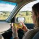 An over-the-shoulder photograph of a passenger in a car holding an iPhone and playing Stardew Valley, with a scenic, sunny country road and farm fields visible through the vehicle’s windows.