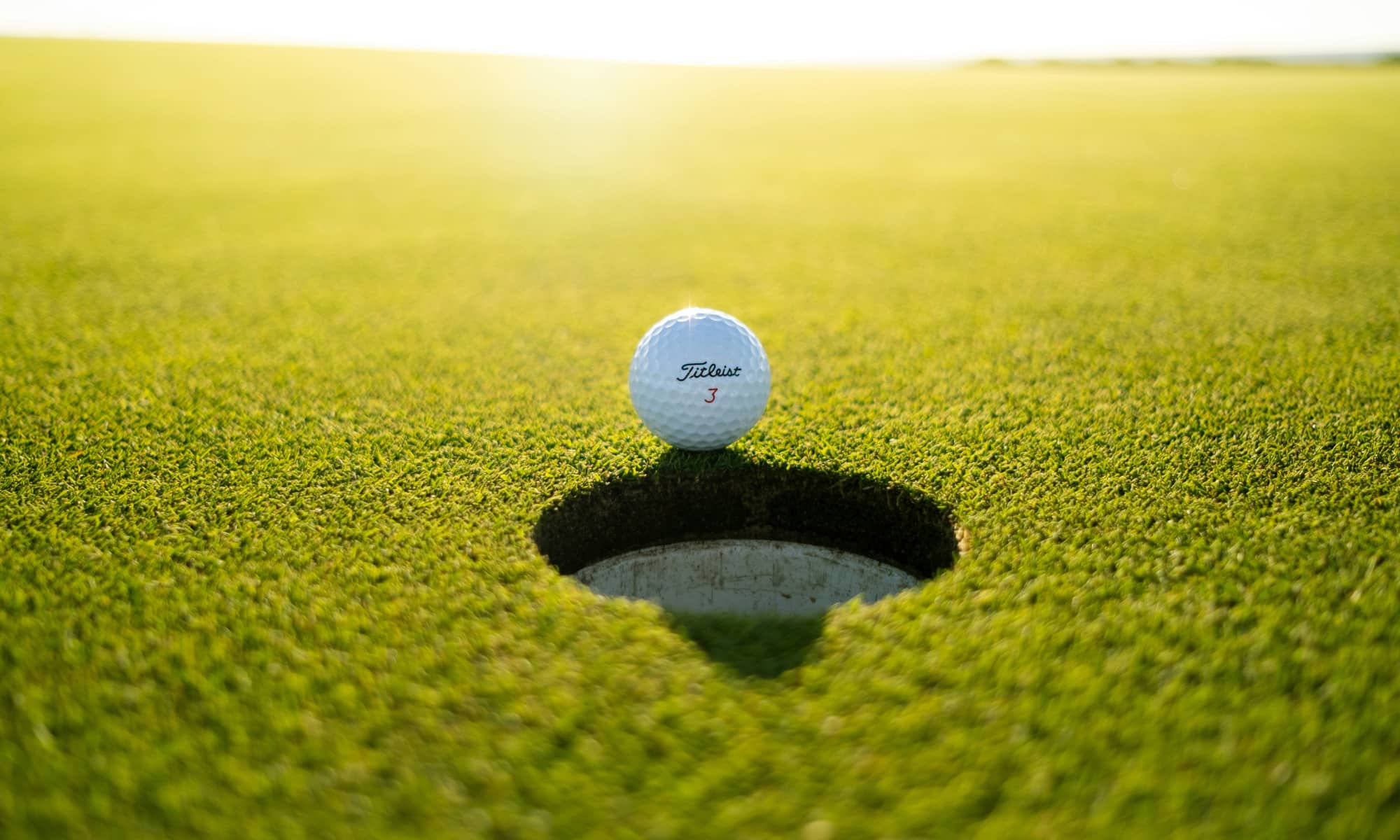 A golf ball on the green sitting at the edge of the hole with the sun in the background.