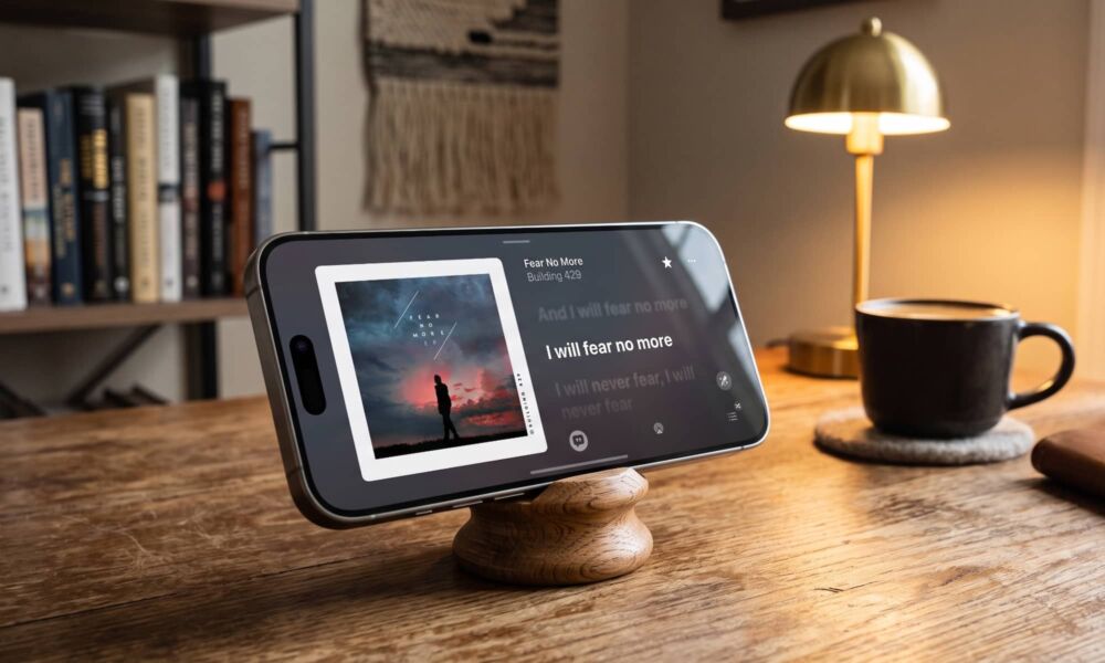 A photo of an iPhone Pro propped on a wooden stand in a cozy home office, displaying the new Apple Music landscape "Now Playing" screen feature coming in iOS 27. The screen shows album art and lyrics for "Fear No More" by Building 429. A coffee mug and lamp are in the background.