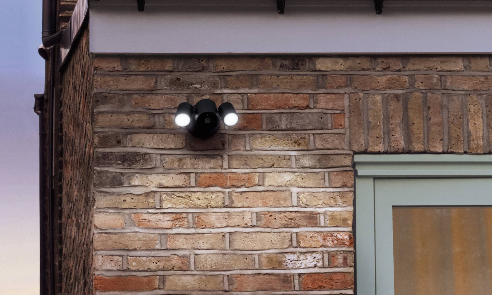 Floodlight Cam in Essence Graphite is mounted on a brick wall above a door, illuminating the area at night.