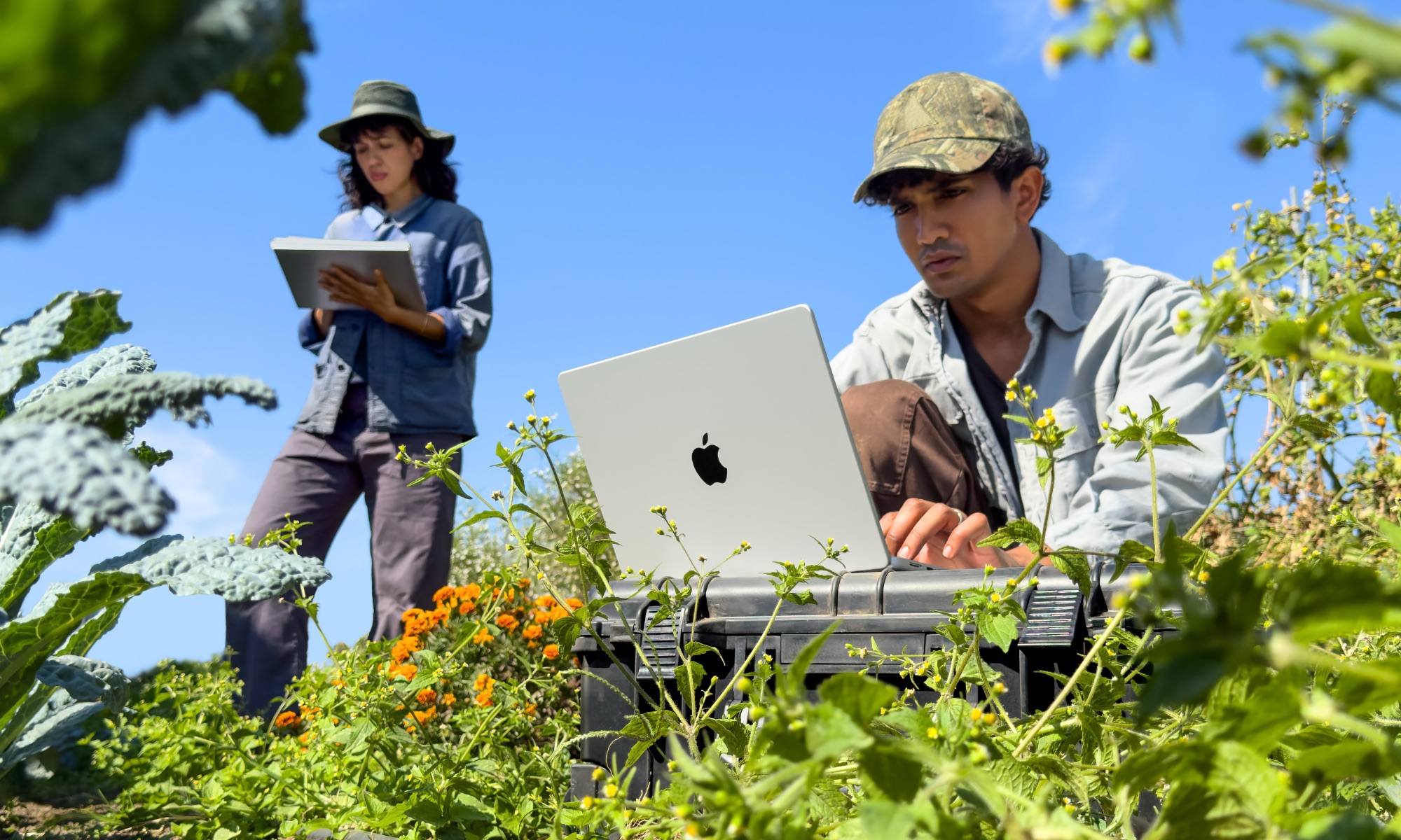 Man using an M6 MacBook Pro in a field while another woman stands in the background taking notes.