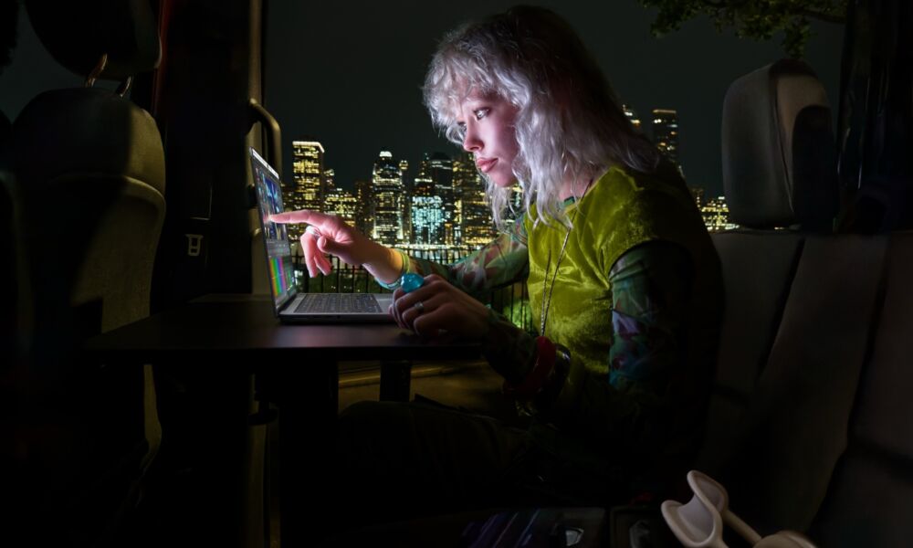 Women in dark hotel room touching the screen on the Apple MacBook Pro with PROLED XDR Display with city skyline in the background.