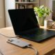 A MacBook Pro and an iPhone resting on a sunlit wooden desk in a home office, connected to braided USB-C charging cables next to a ceramic coffee mug and a small potted plant.