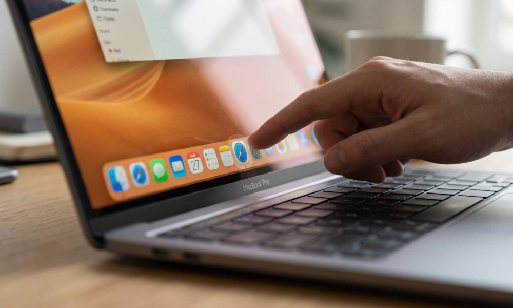 A close-up, side-angled photograph of a hand reaching out to touch the screen of a MacBook Pro resting on a wooden desk. The index finger is pointing toward and lightly touching a Dock icon (like Safari) at the bottom of the display. The macOS Dock, black bezel with the “MacBook Pro” name, and the laptop's keyboard are partially visible under natural indoor lighting, with a shallow depth of field focusing on the interaction point.